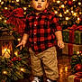 toddler, child, christmas, holiday, festive, lights, presents, lantern, red_shirt, plaid, pants, sneakers, indoors, decorations, pinecones, berries, tree, smiling, standing, floor