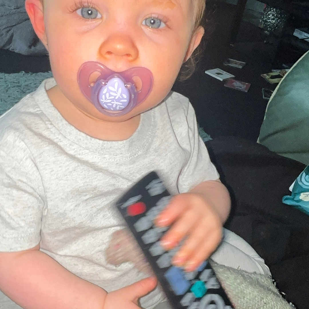 Nala is registered to the contest to win money with this photo: baby_bottle, blanket, blue_eyes, casual, child, clothing, curious, face, furniture, hand, home, indoor, infant, pacifier, person, remote_control, sitting, toddler, toy, young_child