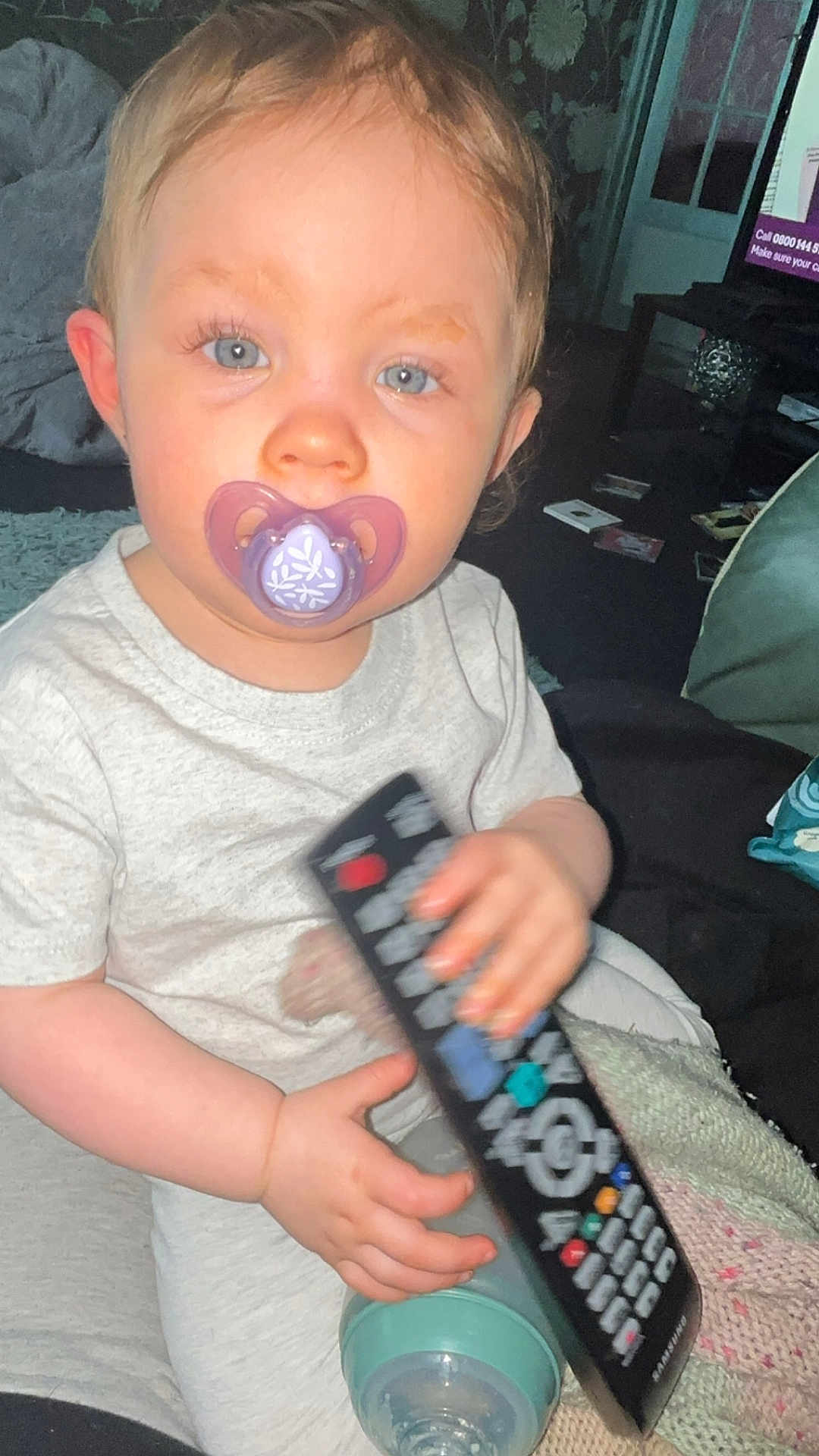 Nala is registered to the contest to win money with this photo: toddler, child, pacifier, remote_control, baby_bottle, indoor, blue_eyes, clothing, person, sitting, hand, face, curious, casual, home, blanket, furniture, toy, young_child, infant