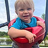 River is registered to the contest to win money with this photo: toddler, child, swing, playground, outdoor, park, smiling, blonde_hair, blue_shirt, red_swing, grass, trees, sky, pond, happy, hands, shorts, daytime, person, cute