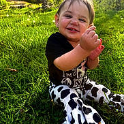 Kamariah is registered to the contest to win money with this photo: toddler, child, grass, outdoor, smiling, black_shirt, cow_print_pants, greenery, nature, sunlight, happy, playing, baby, face, person, grass_field, dandelion, casual_clothing, cute, sitting