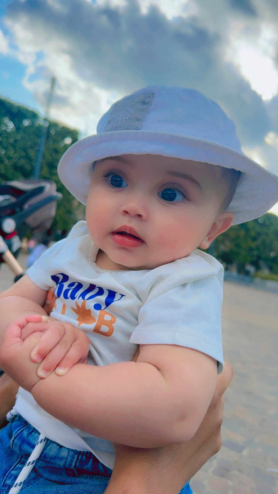 Alseny-Arthur a rejoint le concours — aidez-le/la à gagner de superbes lots ! baby, child, hat, white_hat, tshirt, blue_jeans, outdoor, sky, clouds, hand, person, cute, portrait, holding, greenery, daylight, expression, infant, casual_clothing, closeup