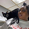 cat, black_cat, woman, person, portrait, selfie, indoor, kitchen, shoulder, curly_hair, black_fur, golden_eyes, pet, companion, close_up, casual, shirt, ceiling_tiles, home, looking_at_camera