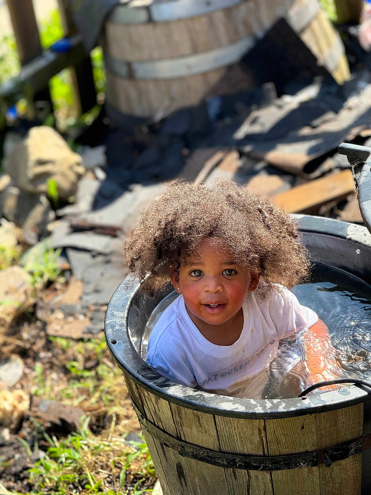 Karti is registered to the contest to win money with this photo: child, water, bucket, outdoor, sunlight, curly_hair, happy, splashing, wooden_bucket, nature, grass, dirt, play, toddler, casual_clothing, summer, daylight, person, face, smiling