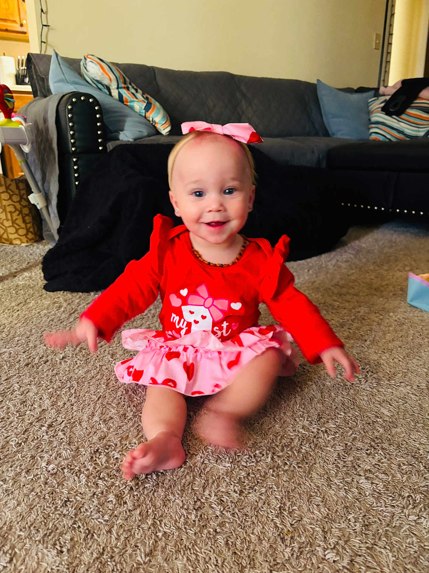 Ember is registered to the contest to win money with this photo: baby, toddler, child, sitting, smile, red_dress, heart_pattern, pink_bow, headband, necklace, carpet, living_room, couch, throw_blanket, decorative_pillows, toy, baby_seat, barefoot, motion_blur, portrait