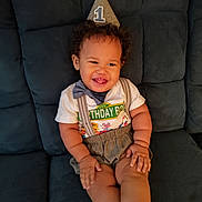 Jeremiah is registered to the contest to win money with this photo: baby, birthday_hat, party_hat, smiling, child, toddler, suspenders, bow_tie, shorts, sitting, couch, happy, curly_hair, indoors, celebration, clothing, face, person, one_year_old, cute
