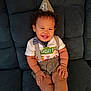 baby, birthday_hat, party_hat, smiling, child, toddler, suspenders, bow_tie, shorts, sitting, couch, happy, curly_hair, indoors, celebration, clothing, face, person, one_year_old, cute