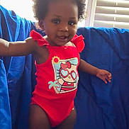 Jaycee is registered to the contest to win money with this photo: toddler, child, swimsuit, red_clothing, blue_bow, curly_hair, standing, indoors, window, blinds, blue_backdrop, smiling, cute, skin, person, happy, young_child, playful, portrait, sunlight