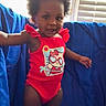 toddler, child, swimsuit, red_clothing, blue_bow, curly_hair, standing, indoors, window, blinds, blue_backdrop, smiling, cute, skin, person, happy, young_child, playful, portrait, sunlight