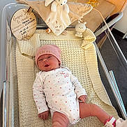 Ariana a rejoint le concours — aidez-le/la à gagner de superbes lots ! baby, baby_clothes, baby_girl, bassinet, blanket, cute_expression, face, hospital, hospital_band, indoor, infant, knitted_blanket, lying_down, newborn, onesie, person, pink_hat, soft_toy, toy, wooden_plaque
