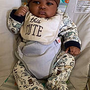 Jakari is registered to the contest to win money with this photo: baby, infant, child, onesie, hat, bib, baby_seat, patterned_clothing, animal_print, indoor, floor, serious_expression, cute, chubby_cheeks, sitting, face, hand, footed_pajamas, colorful_background, closeup