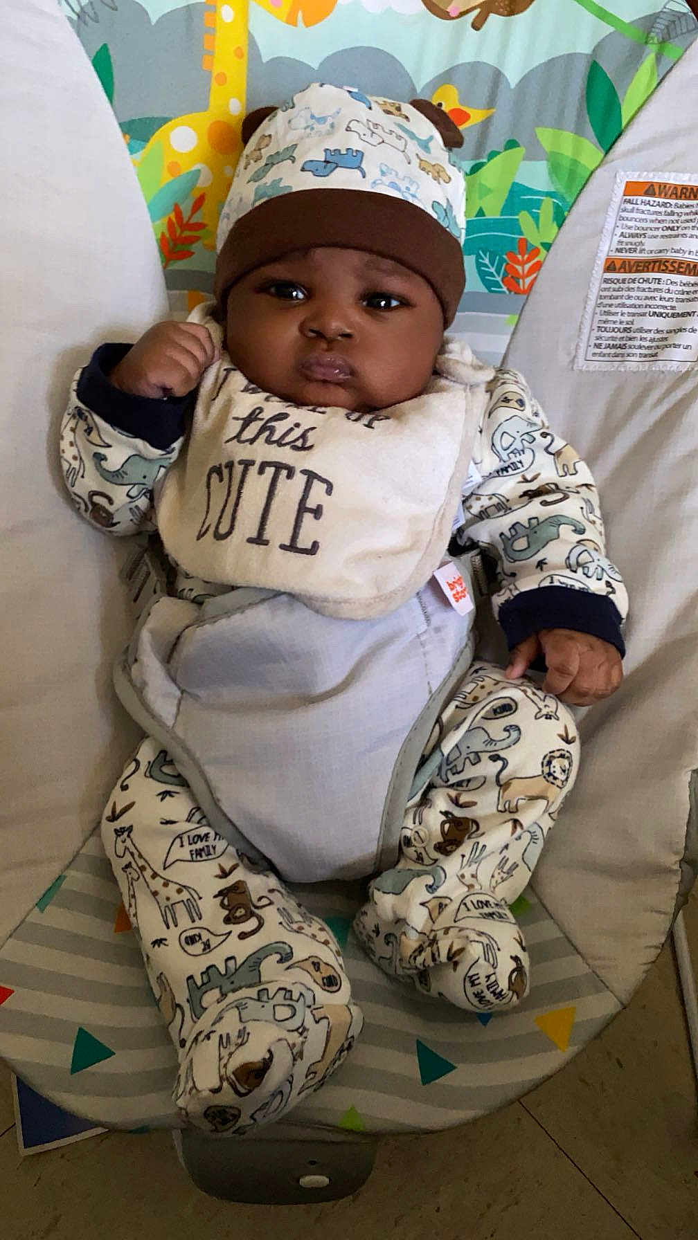 Jakari is registered to the contest to win money with this photo: baby, infant, child, onesie, hat, bib, baby_seat, patterned_clothing, animal_print, indoor, floor, serious_expression, cute, chubby_cheeks, sitting, face, hand, footed_pajamas, colorful_background, closeup