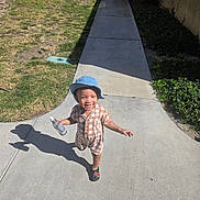 Miles is registered to the contest to win money with this photo: child, toddler, bucket_hat, smile, sidewalk, shadow, spray_bottle, checkered_outfit, shoes, grass, tree, hedge, building, pathway, sunny, outdoor, playful, portrait, concrete, urban_residential