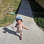 child, toddler, bucket_hat, smile, sidewalk, shadow, spray_bottle, checkered_outfit, shoes, grass, tree, hedge, building, pathway, sunny, outdoor, playful, portrait, concrete, urban_residential