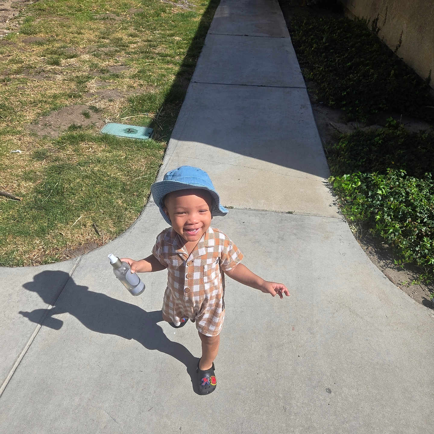 Miles is registered to the contest to win money with this photo: bucket_hat, building, checkered_outfit, child, concrete, grass, hedge, outdoor, pathway, playful, portrait, shadow, shoes, sidewalk, smile, spray_bottle, sunny, toddler, tree, urban_residential