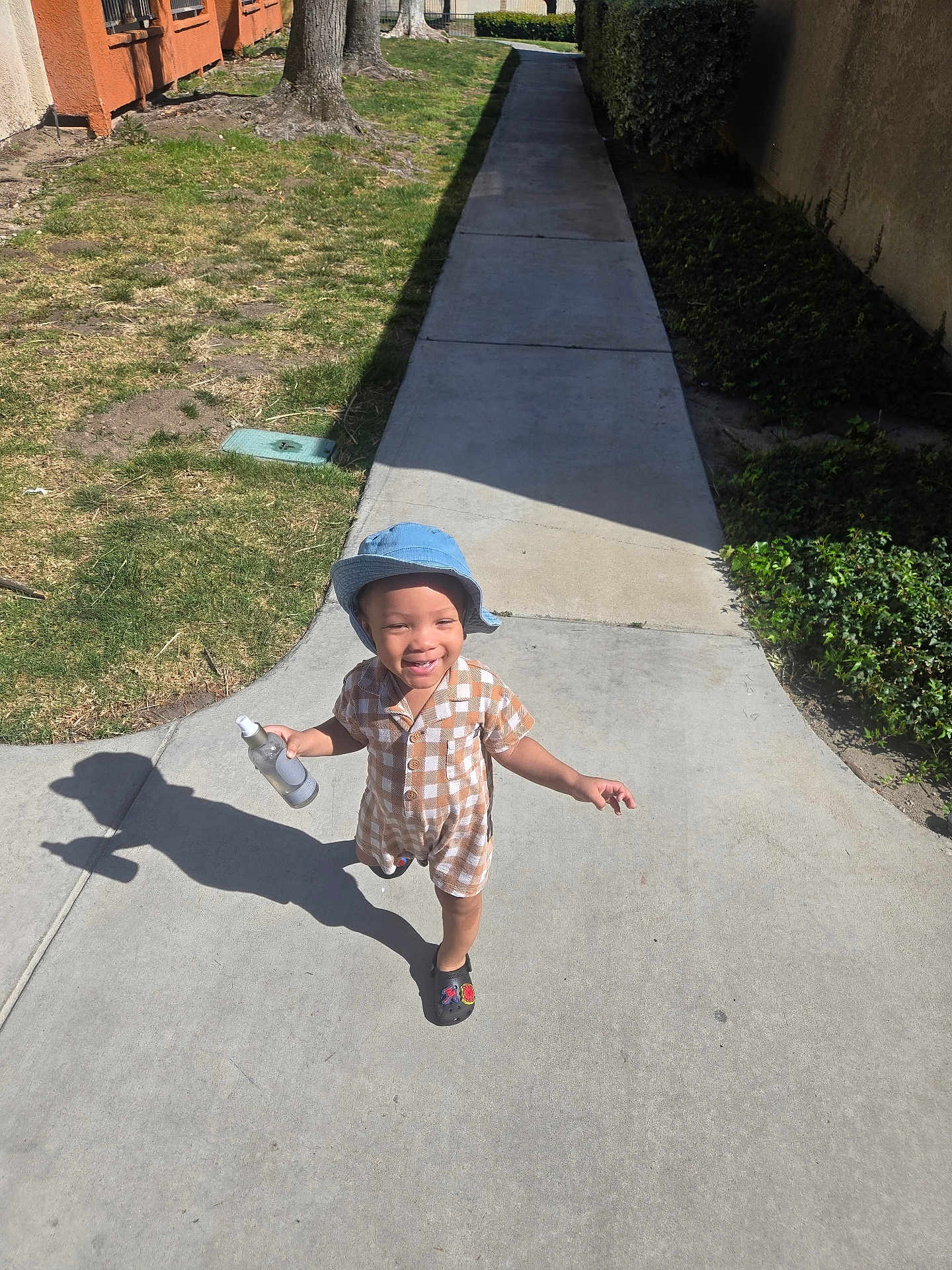 Miles is registered to the contest to win money with this photo: child, toddler, bucket_hat, smile, sidewalk, shadow, spray_bottle, checkered_outfit, shoes, grass, tree, hedge, building, pathway, sunny, outdoor, playful, portrait, concrete, urban_residential