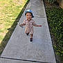 child, toddler, smiling, denim_hat, checkered_outfit, bottle, walking, sidewalk, concrete, grass, lawn, tree, shrub, greenery, shadow, shoes, sandals, outdoors, portrait, candid