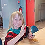 blonde_hair, blue_chair, butterfly_decoration, casual_clothing, child, fire_extinguisher, floor_tiles, hand_gesture, indoor, light, peace_sign, person, playful, portrait, red_door, smiling, table, toy, wall, young