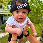 Zakai joined the competition — help win amazing prizes! baby, child, infant, cap, hat, black_cap, white_shirt, outdoor, greenery, palm_leaves, hands, person, grass, house, smiling, cute, portrait, young_child, playing, nature