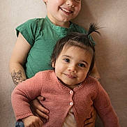 Arya a rejoint le concours — aidez-le/la à gagner de superbes lots ! child, smiling, portrait, cute, happy, sibling, indoor, casual_clothing, green_shirt, pink_cardigan, baby_hair, holding, toddler, young_child, face, person, warm_lighting, tattoo, jeans, background