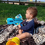Nino participe au concours pour gagner de l'argent avec cette photo : toddler, child, water_gun, yellow_bucket, sand_toys, blanket, grass, outdoor, summer, lake, blue_sky, people_in_background, umbrella, toy, playing, side_view, nature, recreation, sunlight, casual_clothing
