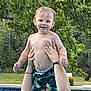 Adrian is registered to the contest to win money with this photo: toddler, child, swimming_pool, water, summer, outdoor, greenery, tree, happy, smiling, person, holding, swim_trunks, bare_chest, adult_hands, nature, sunlight, backyard, playful, fun