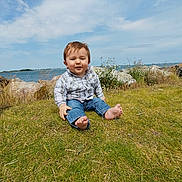 Victor participe au concours pour gagner de l'argent avec cette photo : barefoot, casual_clothing, child, clouds, daytime, grass, greenery, jeans, nature, outdoor, person, plaid_shirt, relaxing, rocks, shoreline, sky, smiling, toddler, water, young_child
