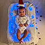 baby, blanket, blue_lights, christmas, cute, decorations, festive, headband, holiday, indoors, infant, lying_down, onesie, pinecones, santa_hat, small_hands, smiling, soft_texture, white_clothing, wide_eyes