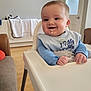 baby, high_chair, smiling, bib, food, face, hands, indoor, living_room, furniture, child, infant, clothing, expression, person, cute, happy, home, wooden_floor, blanket
