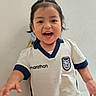 child, toddler, smiling, happy, sports_jersey, white_shirt, navy_trim, indoor, portrait, cute, person, short_hair, dark_hair, cheerful, young_child, face, front_view, casual_clothing, playing, joyful