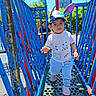 child, toddler, playground, blue_railings, hat, ear_hat, person, adult, outdoor, daylight, smile, walking, clothing, shoes, metal_platform, building, trees, sky, recreation, park