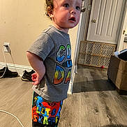 Luca is registered to the contest to win money with this photo: toddler, child, indoor, standing, curly_hair, colorful_clothing, shorts, tshirt, wood_floor, door, baby_gate, shoes, wall, power_outlet, cable, furniture, person, young_child, casual, expression