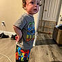 toddler, child, indoor, standing, curly_hair, colorful_clothing, shorts, tshirt, wood_floor, door, baby_gate, shoes, wall, power_outlet, cable, furniture, person, young_child, casual, expression