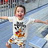 child, smiling, happy, mickey_mouse, pluto, tshirt, shorts, sneakers, bleachers, outdoor, fence, people, joyful, motion, playful, casual_clothing, daylight, portrait, excited, fun