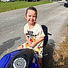 child, smiling, motorcycle, blue_motorcycle, outdoor, street, car, parked_car, clothing, mickey_mouse, happy, person, daylight, asphalt, vehicle, casual, young_child, shorts, tshirt, playful