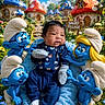 baby, infant, child, smurfs, toys, cartoon_characters, blue_characters, mushroom_houses, fantasy_village, outdoor, garden, flowers, grass, blanket, sitting, portrait, baby_clothing, white_hat, colorful, smile