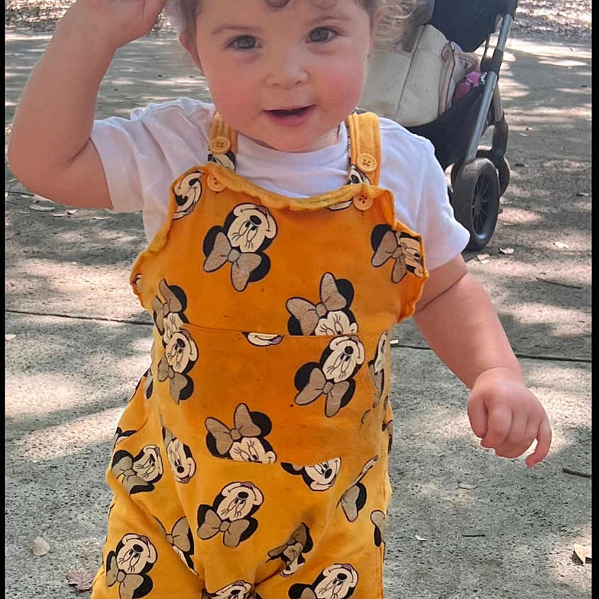 Genesis Hernandez is registered to the contest to win money with this photo: bunny_ears, child, curly_hair, cute, daylight, happy, nature, outdoor, park, person, playful, sidewalk, smiling, sneakers, stroller, sunlight, toddler, trees, white_shirt, yellow_overall