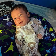 Malakai is registered to the contest to win money with this photo: baby, infant, onesie, animal_print, dinosaur, pillow, bed, blue_wall, smile, face, child, cute, indoors, sleepwear, pacifier, pattern, soft_lighting, portrait, young, cozy