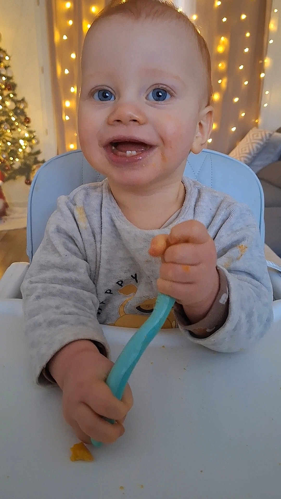 Valentin participe au concours pour gagner de l'argent avec cette photo : toddler, child, high_chair, blue_eyes, spoon, messy_face, smiling, indoor, christmas_tree, fairy_lights, holiday, happy, clothing, furniture, table, portrait, baby, feeding, cute, home