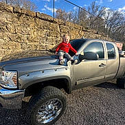 Albert is registered to the contest to win money with this photo: toddler, child, red_sweater, pickup_truck, silver_truck, hood, stone_wall, fence, outdoor, daylight, sky, clouds, gravel, smiling, casual_clothing, vehicle, wheel, reflection, person, nature