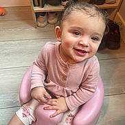 Anaiah is registered to the contest to win money with this photo: toddler, child, baby_seat, pink_clothing, curly_hair, smiling, indoor, wooden_floor, shoe_rack, shoes, orange_ball, floor, cute, happy, person, sitting, portrait, casual, home, playful