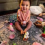 blanket, child, couch, cup, curly_hair, doll, figurine, floor, green_toy, happy, home_interior, indoor, living_room, person, pink_clothing, play, rug, smiling, toddler, toy