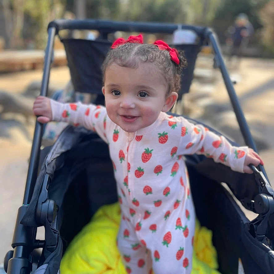 Anaiah joined the competition — help win amazing prizes! baby_clothing, child, curly_hair, daylight, happy, holding, infant, nature, onesie, outdoor, park, person, playful, portrait, red_bow, smiling, strawberry_pattern, stroller, toddler, yellow_blanket