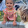 toddler, child, smiling, dog, pet, outdoor, grass, brick_wall, colorful_clothing, playtime, happy, curly_hair, yellow_hair_ties, canopy_frame, nature, cute, person, animal, face, daytime