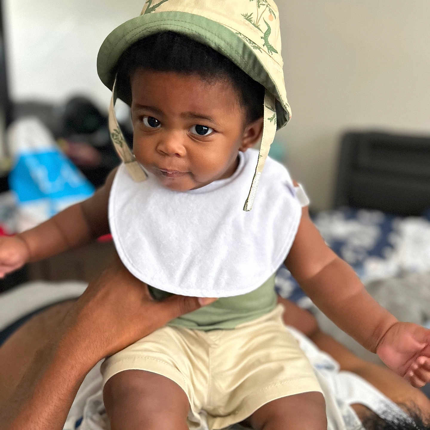 Khiros is registered to the contest to win money with this photo: baby, child, infant, portrait, sitting, hat, bucket_hat, dinosaur_pattern, bib, white_bib, shorts, green_clothing, indoors, home, adult, lap, smiling, wide_eyes, blurred_background, family