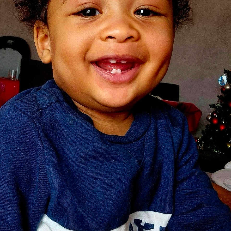 Elio participe au concours pour gagner de l'argent avec cette photo : baby, child, christmas_tree, closeup, cozy, curly_hair, cute, expression, face, furniture, happy, home, indoor, person, portrait, seated, skin, smiling, sweater, toddler