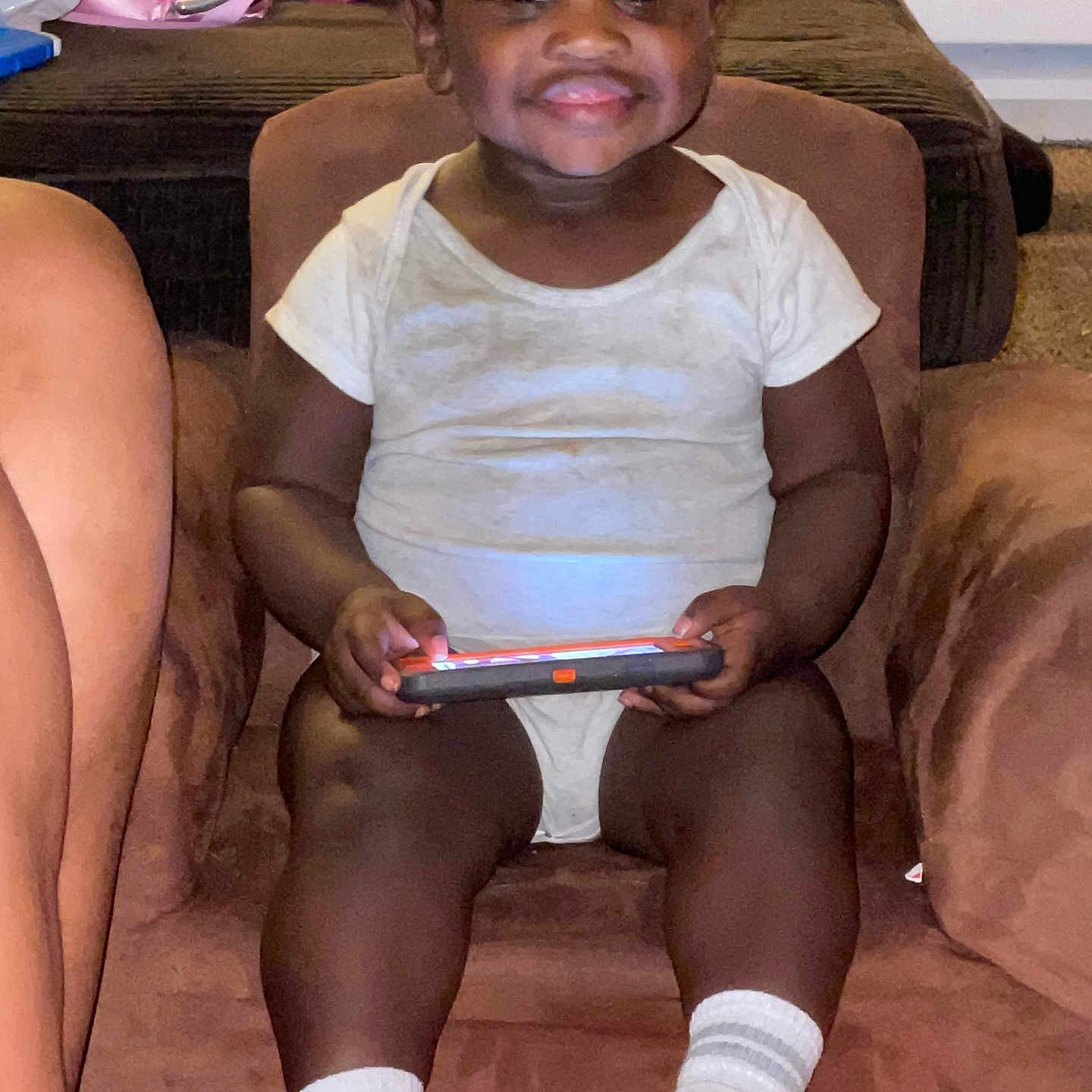 Azkari is registered to the contest to win money with this photo: armchair, brown_chair, carpet, child, cute, feet, furniture, happy, home, indoor, legs, person, play, sitting, smartphone, smiling, toddler, toy_stroller, white_onesie, white_socks