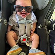 Carter is registered to the contest to win money with this photo: baby, sunglasses, car_seat, smiling, happy, child, seatbelt, casual_shoes, infant, cute, indoor, clothing, person, toddler, funny, headwear, safety, closeup, joyful, accessory