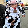 Carter is registered to the contest to win money with this photo: baby, child, costume, cow_costume, plush, outdoor, parking_lot, car, building, person, cute, animal_costume, hood, ears, face, expression, hand, holding, clothing, daytime