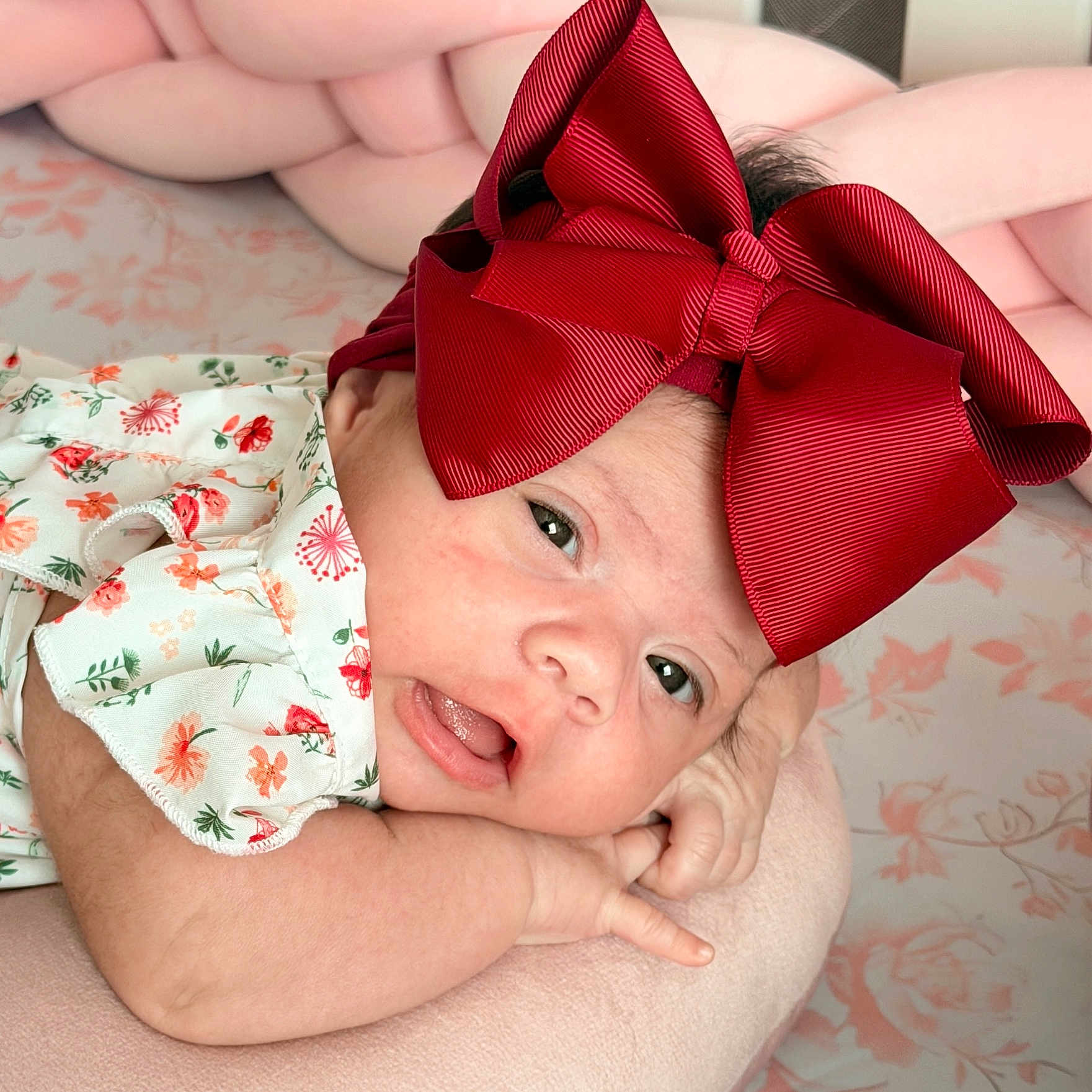 Zoe joined the competition — help win amazing prizes! baby, infant, child, portrait, smiling, happy, red_bow, hair_accessory, floral_clothing, onesie, lying_down, close_up, face, eyes, open_mouth, indoors, blanket, pink_background, adult_arm, family_photo